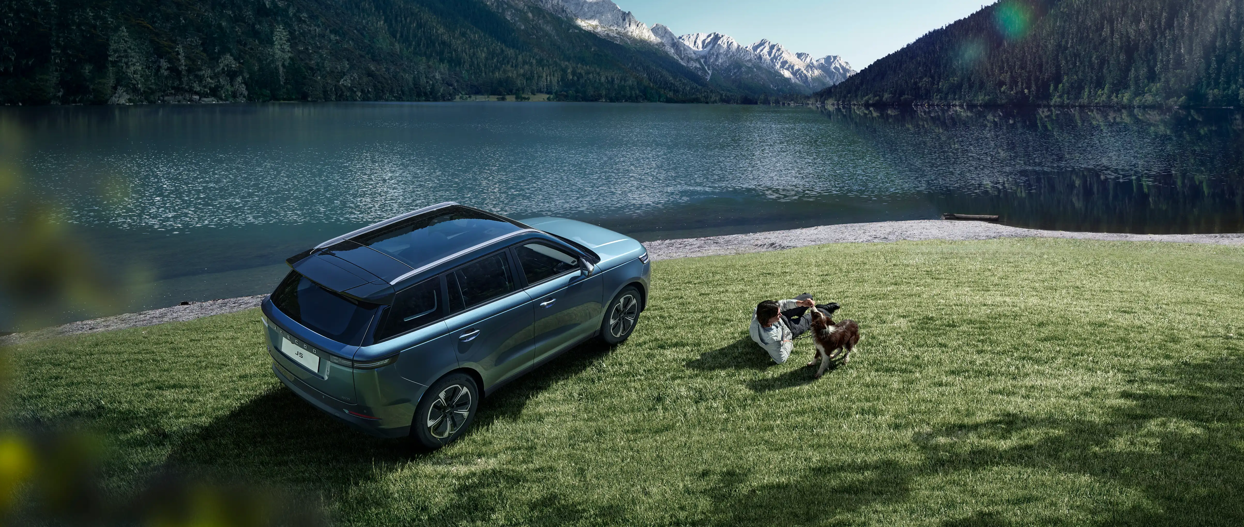 Aerial view of a blue JAECOO 5 parked on green grass by a mountain lake; a person lying on the grass interacts playfully with a dog next to the vehicle.