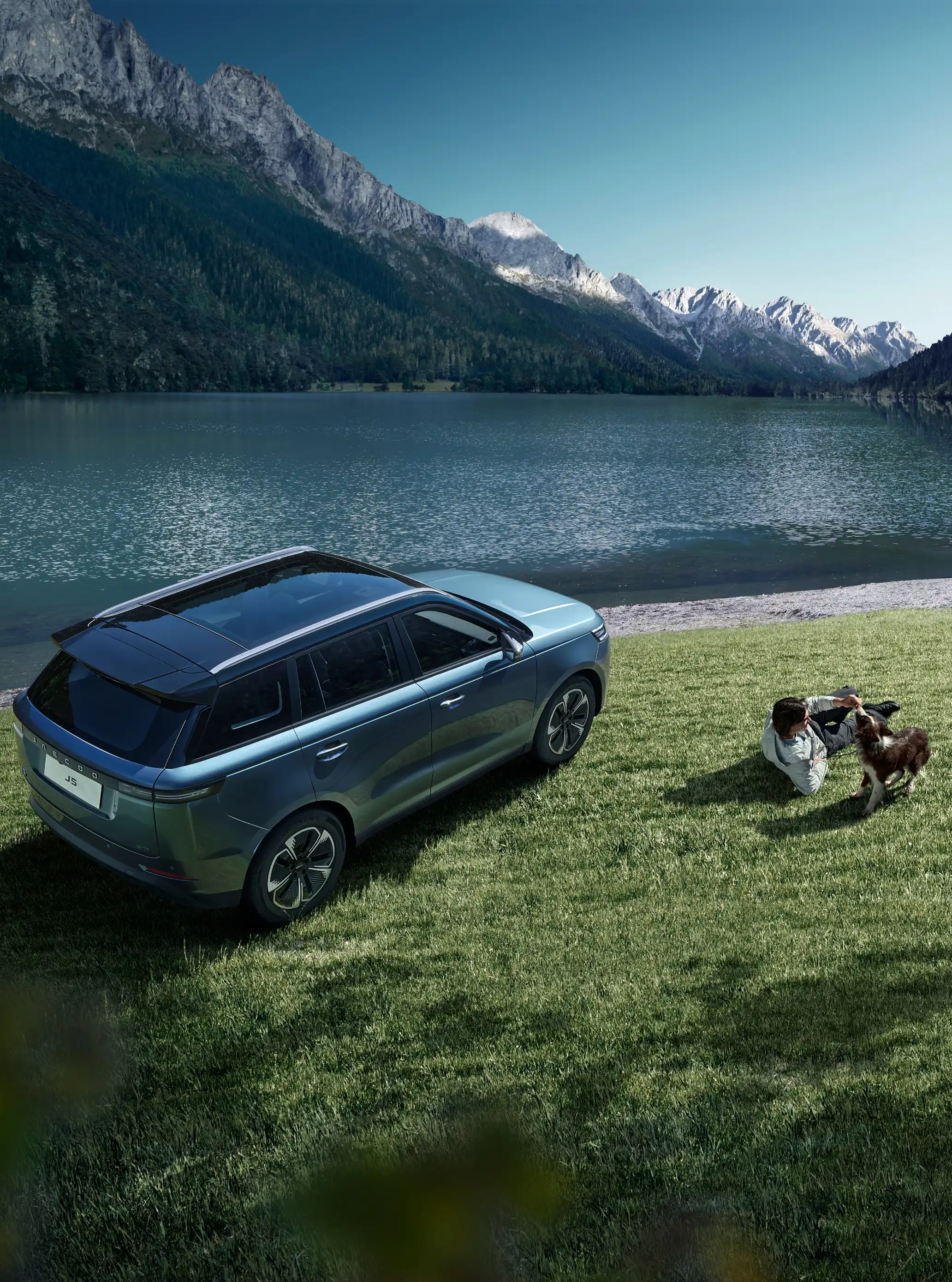Aerial view of a blue JAECOO 5 parked on green grass by a mountain lake; a person lying on the grass interacts playfully with a dog next to the vehicle.