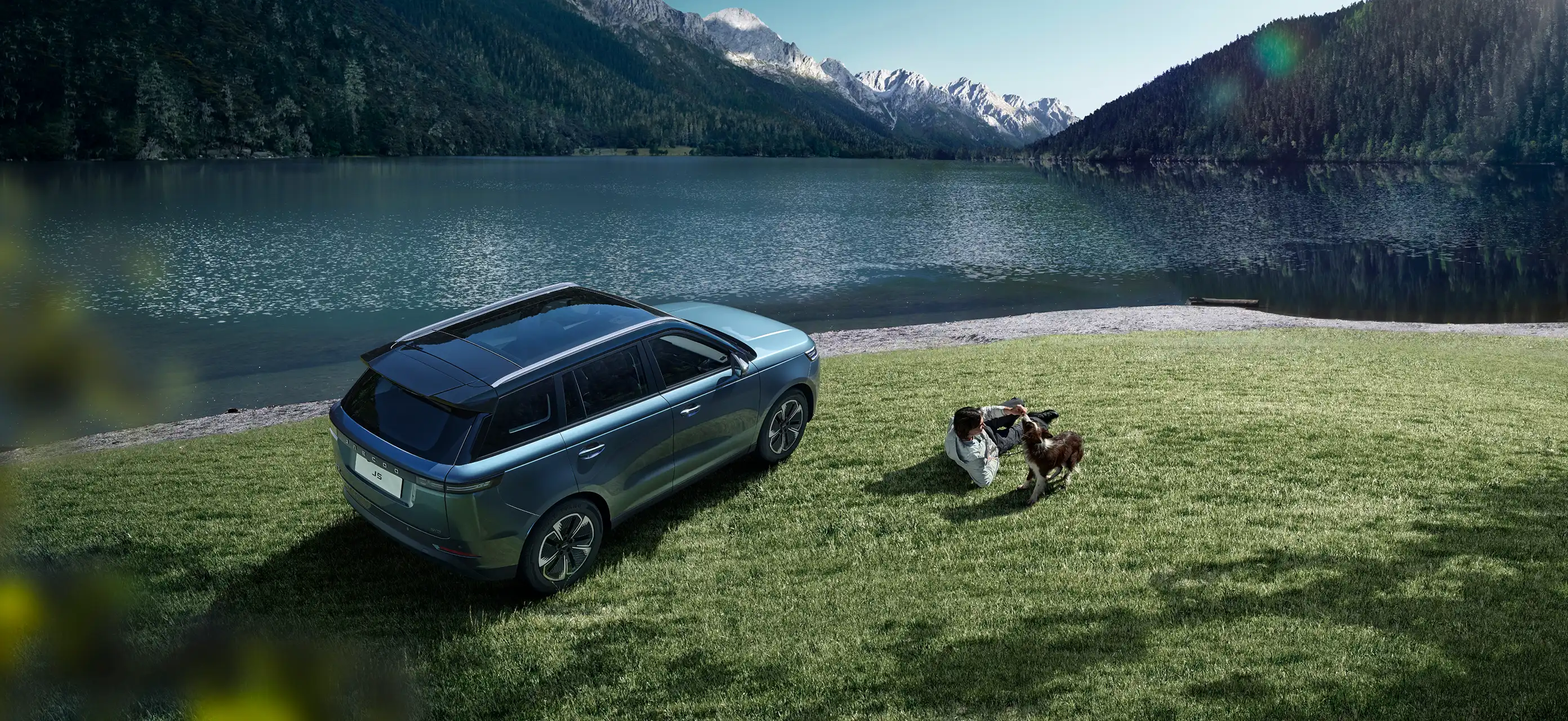 Aerial view of a blue JAECOO 5 parked on green grass by a mountain lake; a person lying on the grass interacts playfully with a dog next to the vehicle.