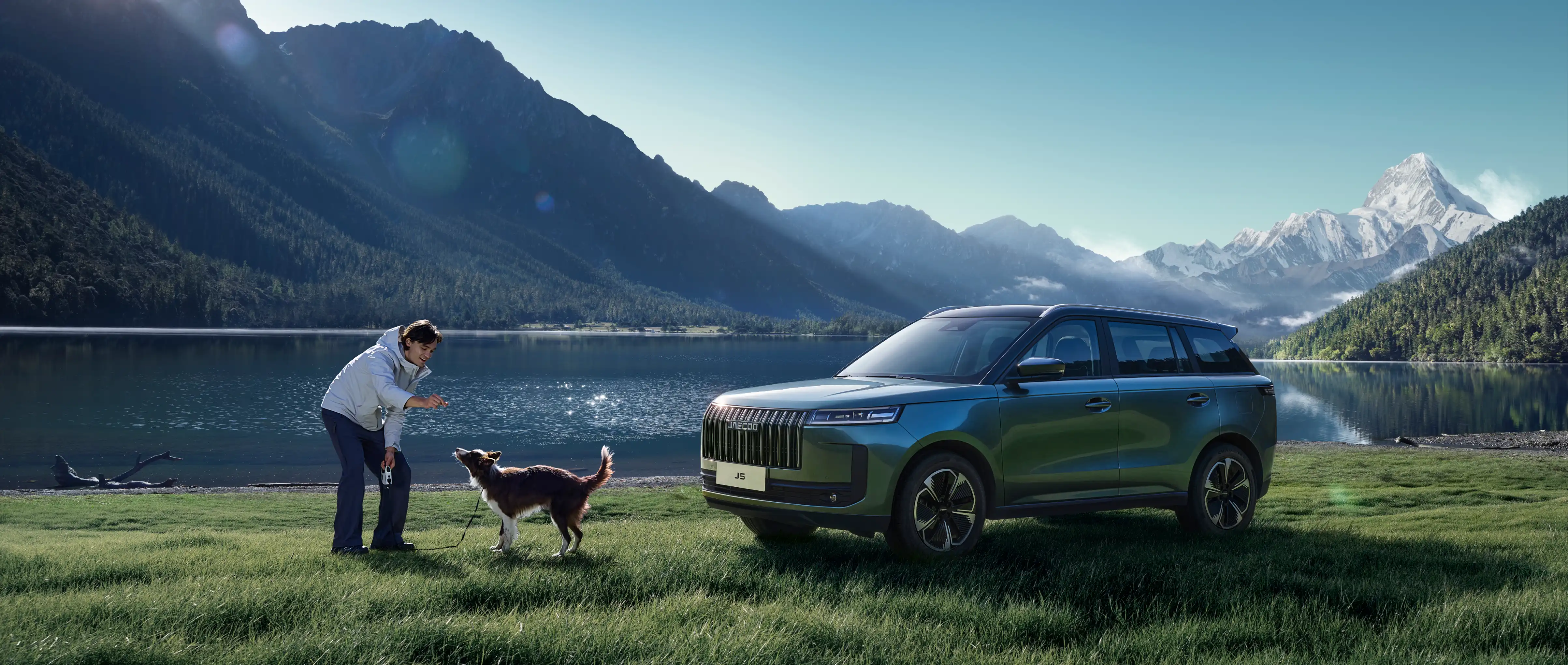 Front three-quarter view of a blue JAECOO 5 parked on a grassy lakeside with snow-capped peaks in the distance; a person bends down to play with a dog.