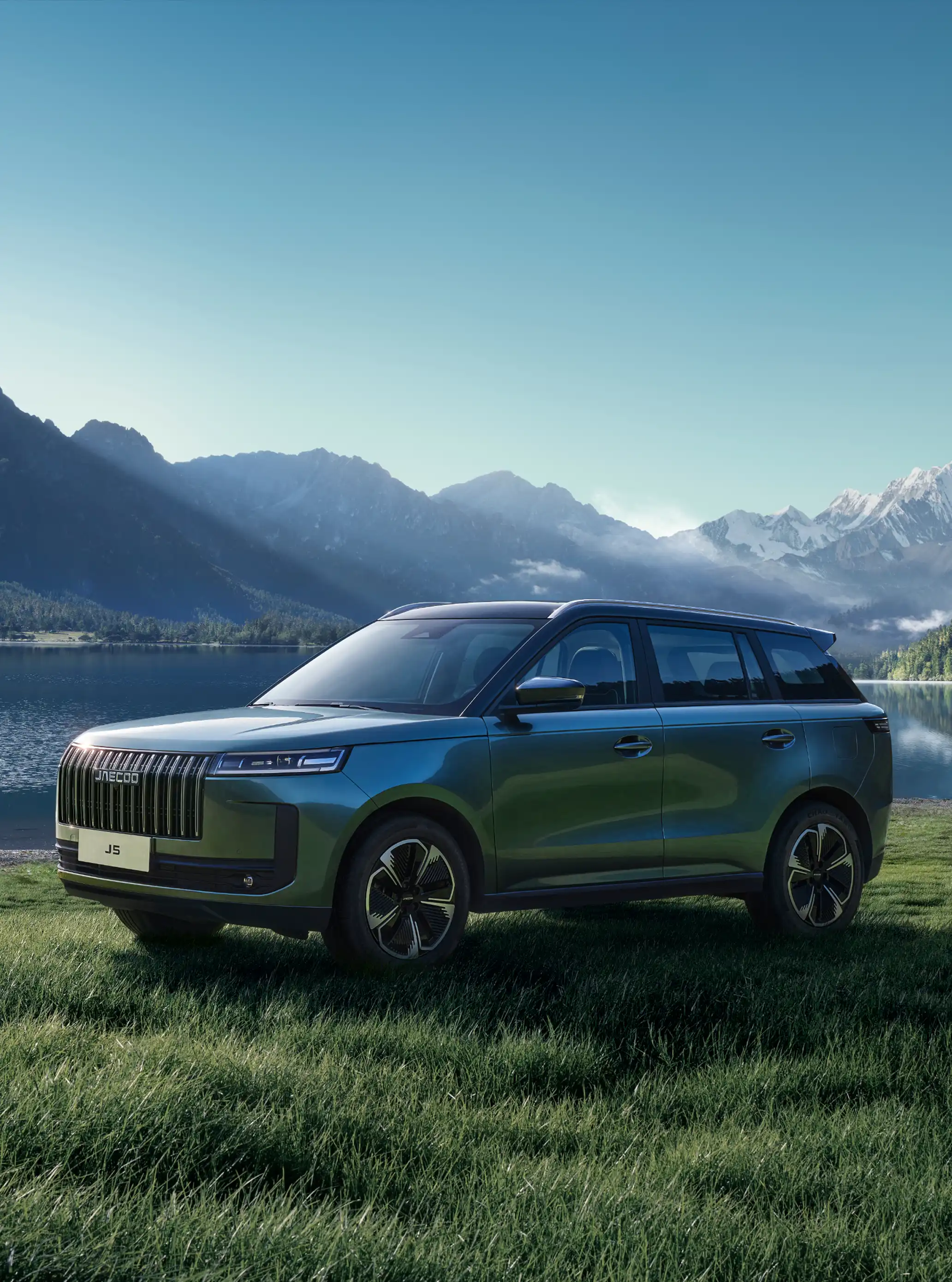 Front three-quarter view of a blue JAECOO 5 parked on a grassy lakeside with snow-capped peaks in the distance; a person bends down to play with a dog.