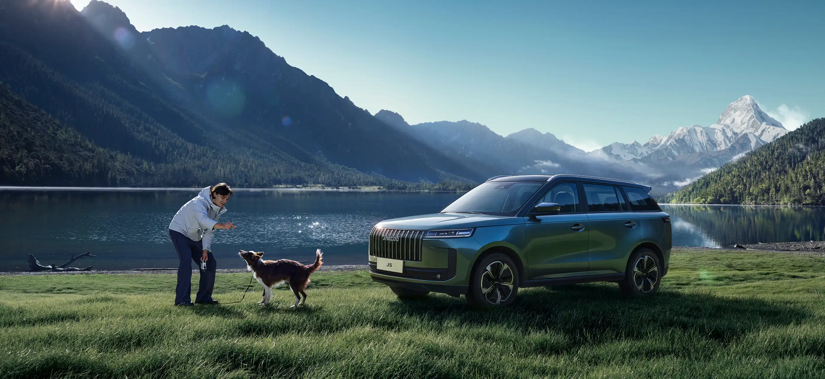 Front three-quarter view of a blue JAECOO 5 parked on a grassy lakeside with snow-capped peaks in the distance; a person bends down to play with a dog.