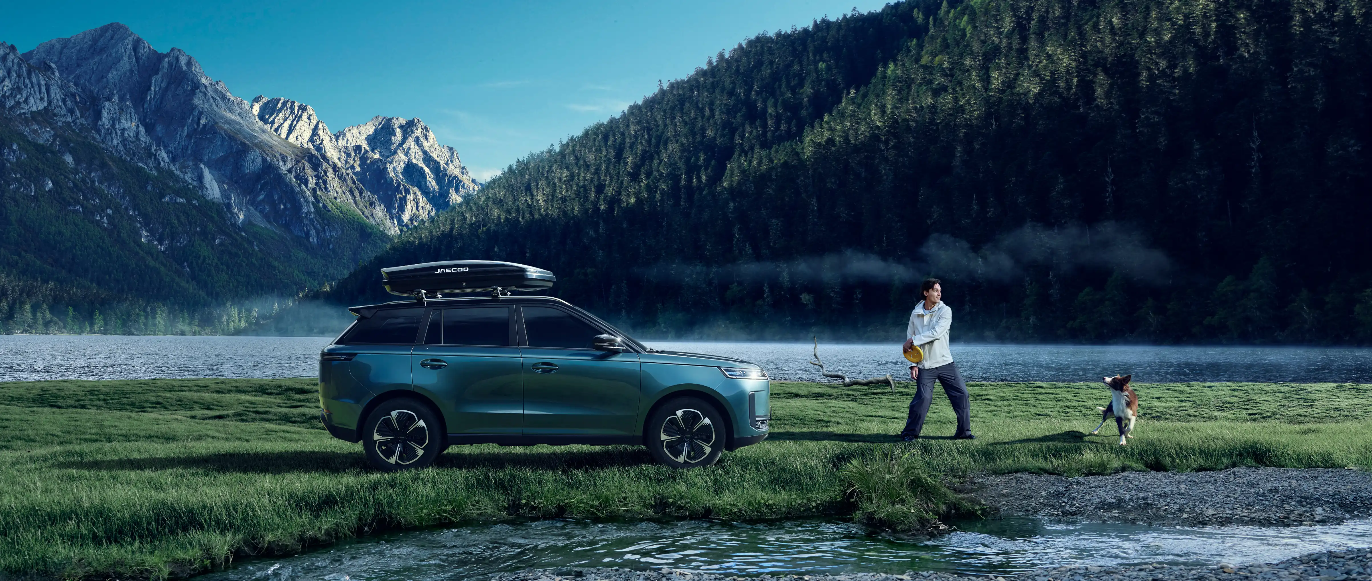 Blue JAECOO 5 with a roof box parked on a misty lakeshore in front of a pine-covered mountain; a person standing with a frisbee plays with a dog beside the water.