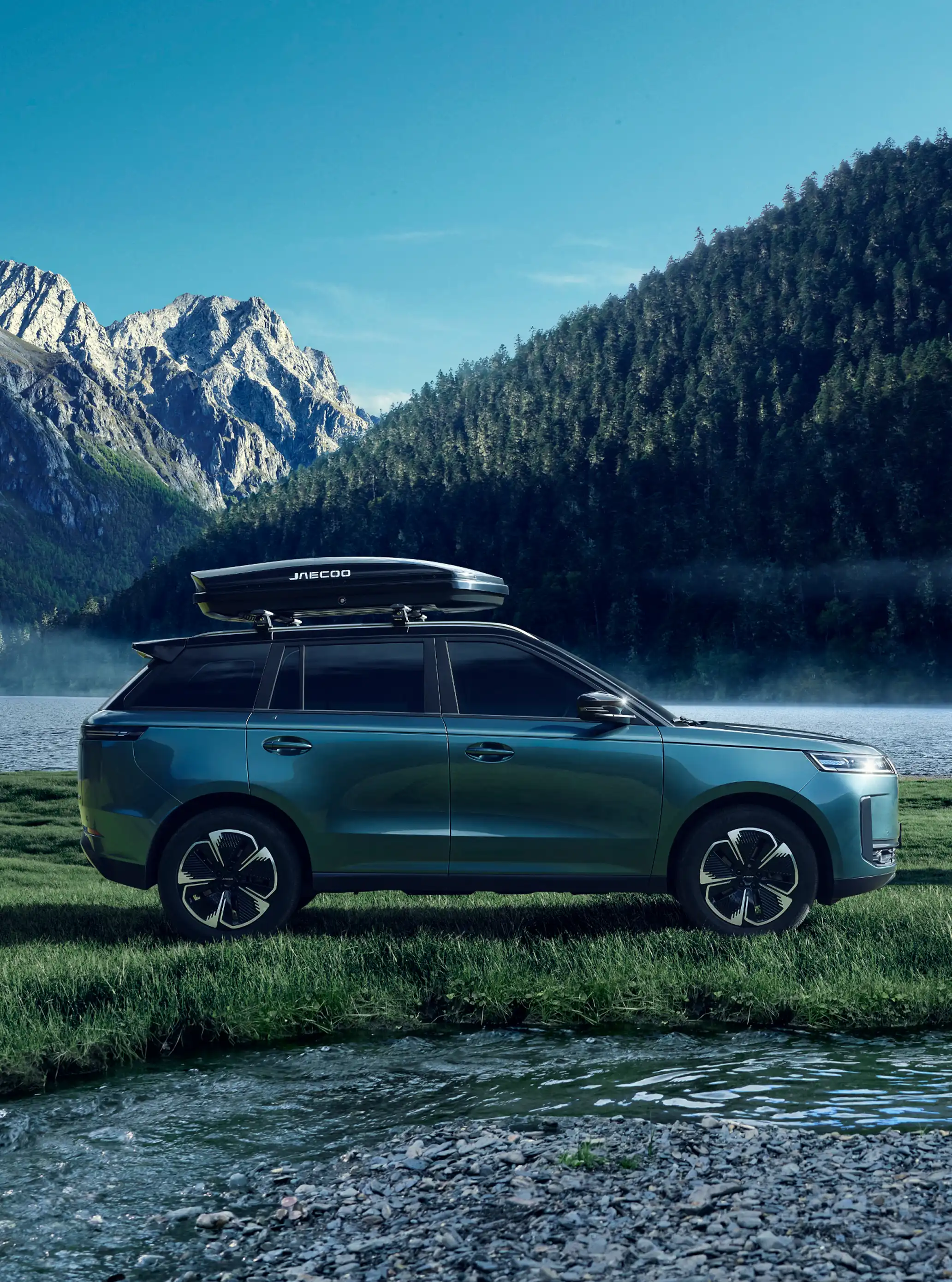 Blue JAECOO 5 with a roof box parked on a misty lakeshore in front of a pine-covered mountain; a person standing with a frisbee plays with a dog beside the water.
