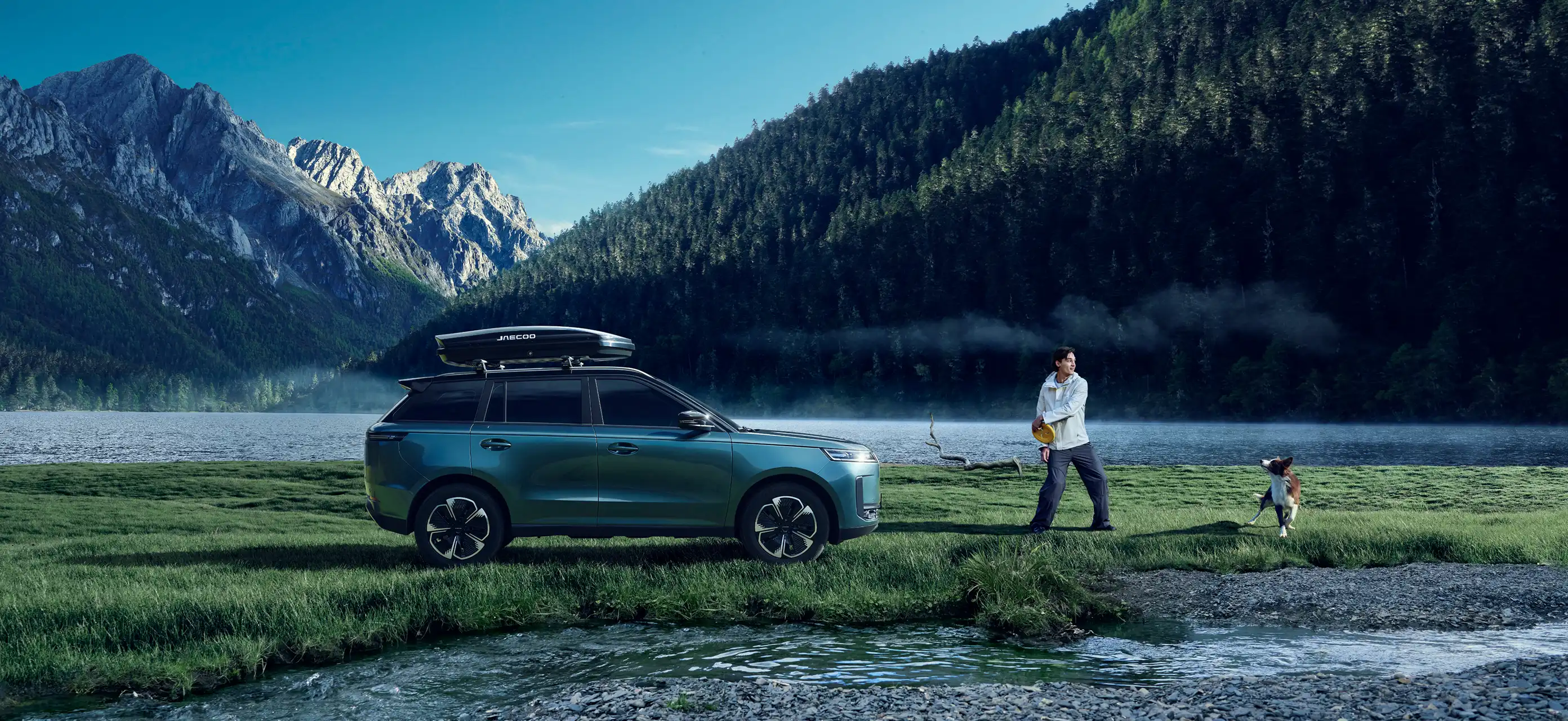 Blue JAECOO 5 with a roof box parked on a misty lakeshore in front of a pine-covered mountain; a person standing with a frisbee plays with a dog beside the water.