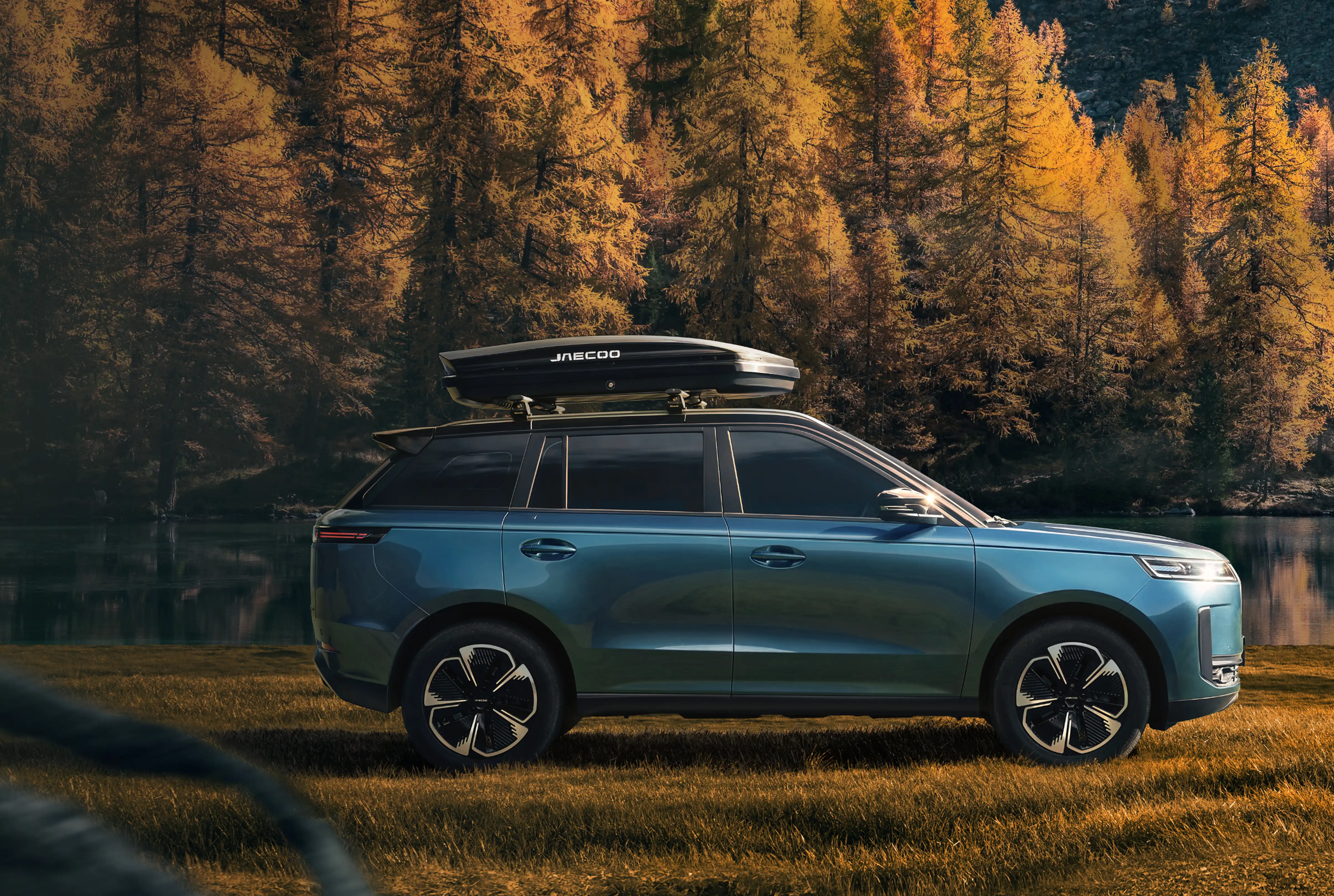 Blue JAECOO 5 SUV with a roof box parked on an autumn lakeshore in a mountain forest, with a person walking a dog alongside it.